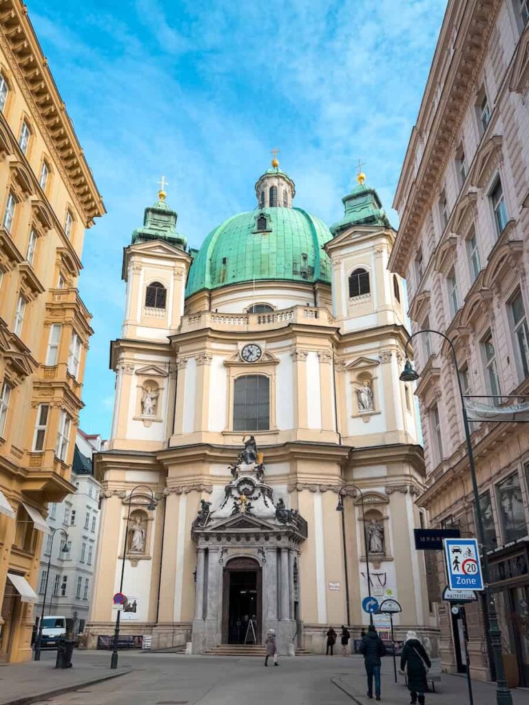 Exterior view of St. Peter’s Church (Peterskirche) in Vienna, Austria, featuring its iconic green copper dome, Baroque architecture, and ornate façade with statues, nestled between historic buildings on a quiet pedestrian street under a blue sky.