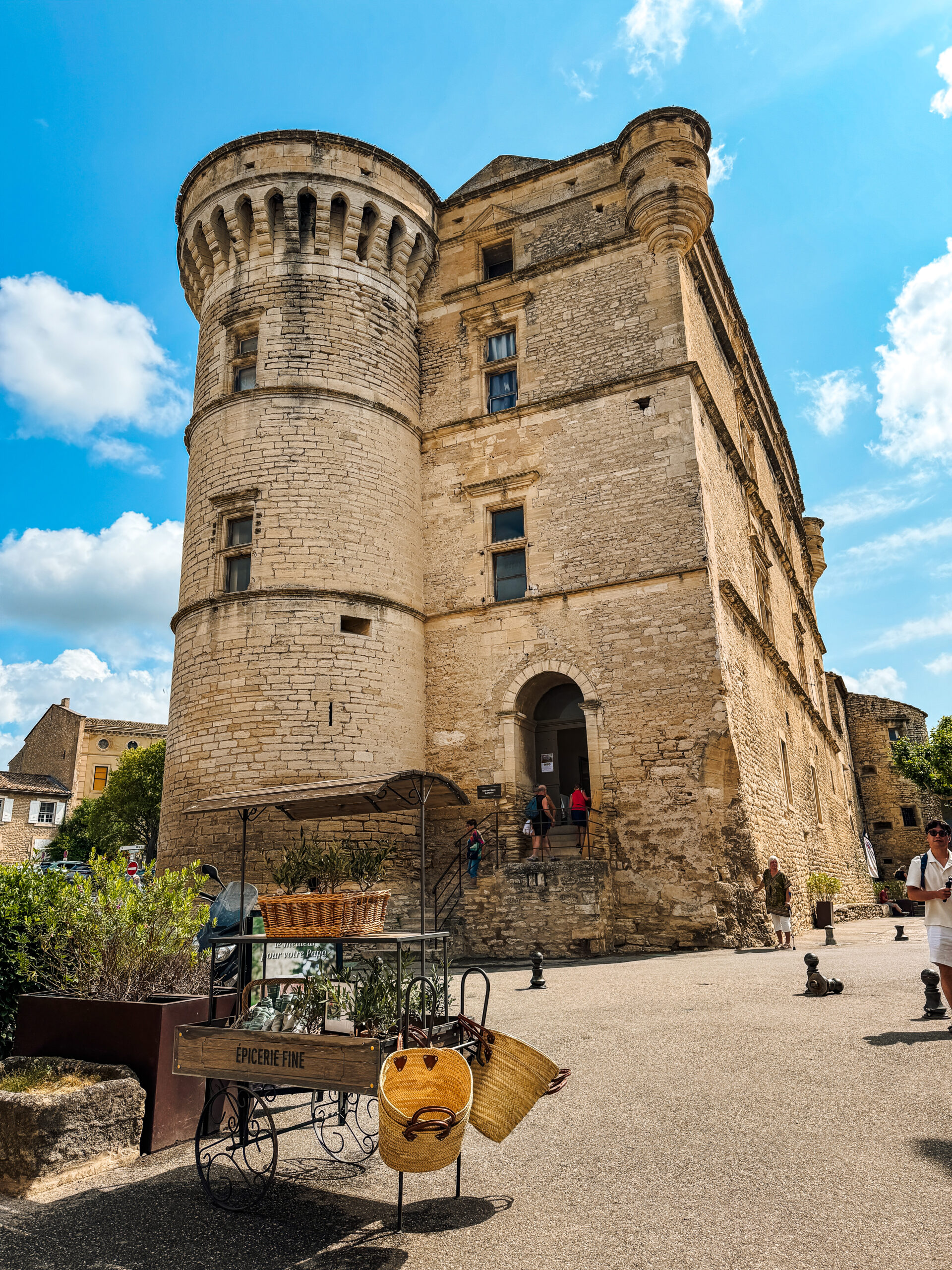 A medieval stone fortress in Gordes, France, with a round tower against a blue Provençal sky, baskets and olive trees in the foreground.