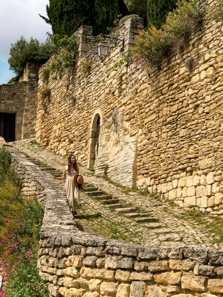 Woman in a flowy dress holding a straw hat, walking along a stone stairway surrounded by golden village walls in Gordes France. 