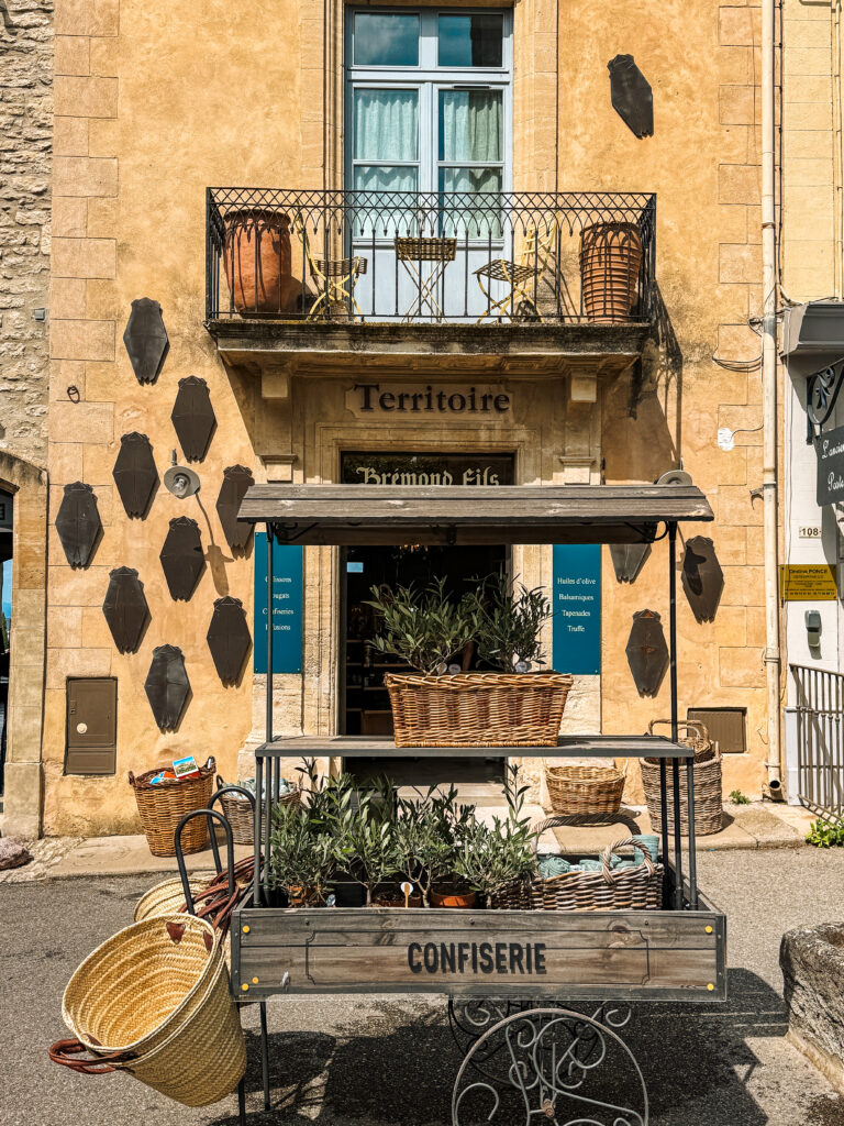 A charming shopfront in Gordes with a cart of olive plants, wicker baskets, and confiserie sign under pastel shutters.