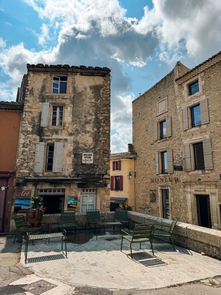 Rustic stone houses with pastel shutters surround a small square with green chairs under a dramatic Provençal sky.