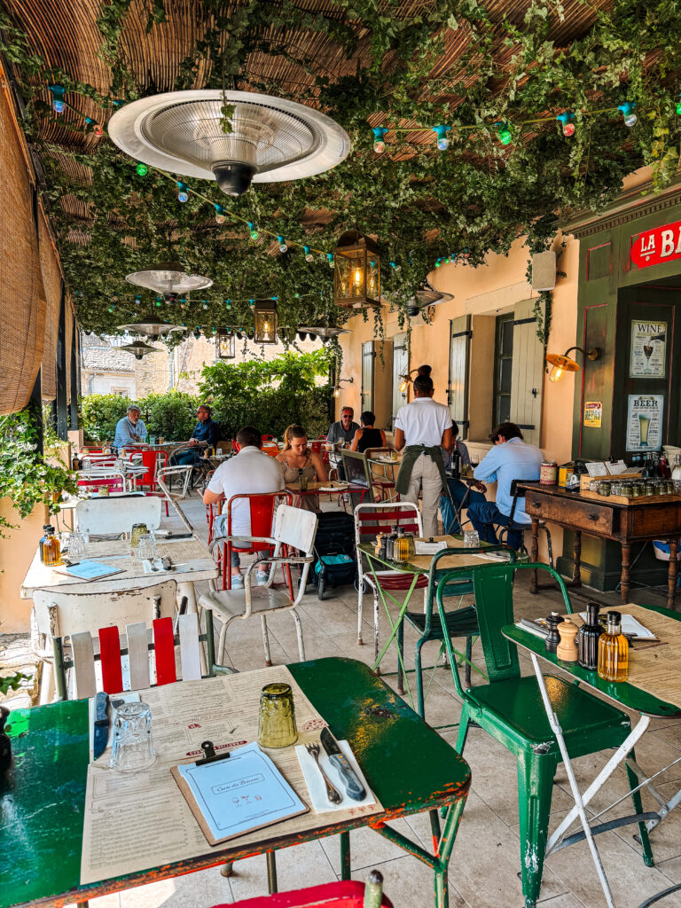 Outdoor dining with colorful metal chairs, leafy overhead canopy, and Provençal charm in Gordes, France.