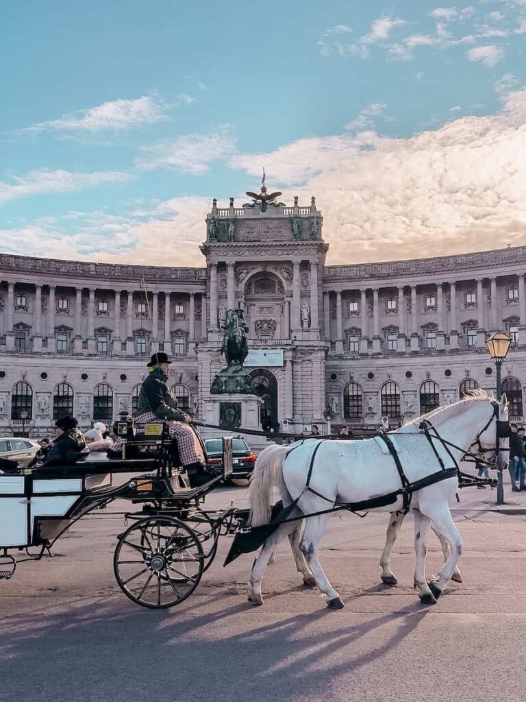 Horse-drawn carriage passing in front of the grand Hofburg Palace in Vienna, Austria, with a statue and ornate architecture in the background under a pastel-colored sky.