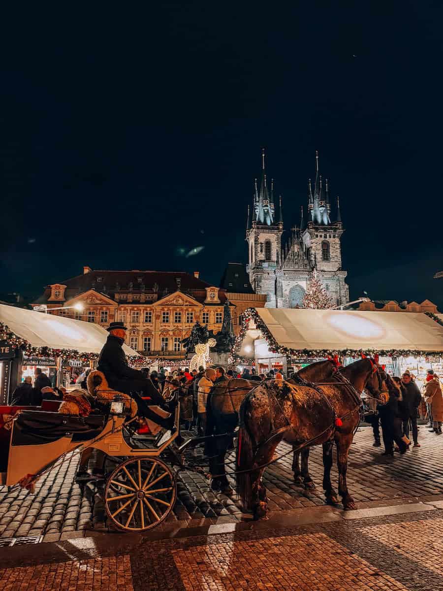 Horse-drawn carriage in front of festive Christmas market stalls in Prague’s Old Town Square at night, with the Church of Our Lady before Týn illuminated in the background and crowds enjoying the holiday atmosphere during Christmas markets in Prague