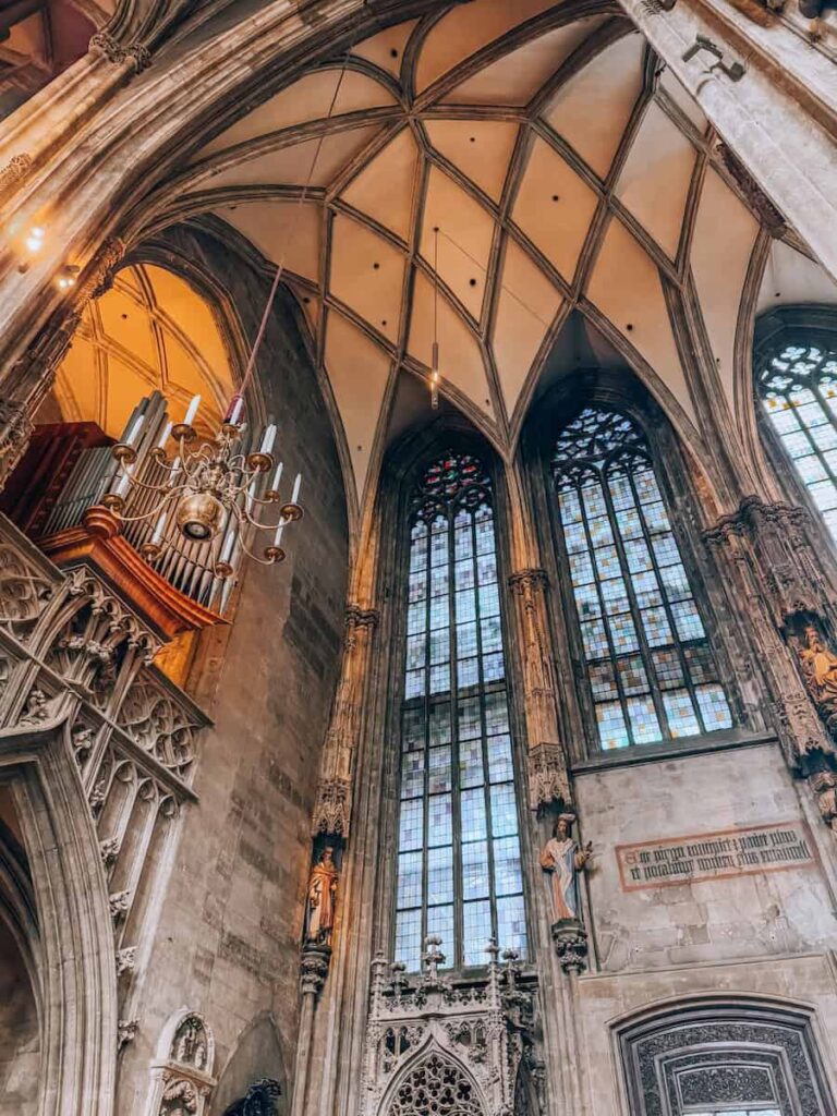 Detailed view of the vaulted ceiling and tall stained glass windows inside St. Stephen’s Cathedral in Vienna. The Gothic architecture features ribbed arches, ornate carvings, statues of saints, and a glimpse of the cathedral’s grand pipe organ.