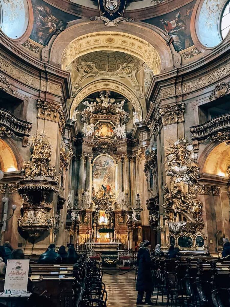 Interior of St. Peter’s Church in Vienna, featuring an opulent Baroque altar adorned with gilded sculptures, dramatic paintings, intricate columns, and angelic figures. Visitors are seen quietly admiring the ornate details beneath the soaring vaulted ceiling.