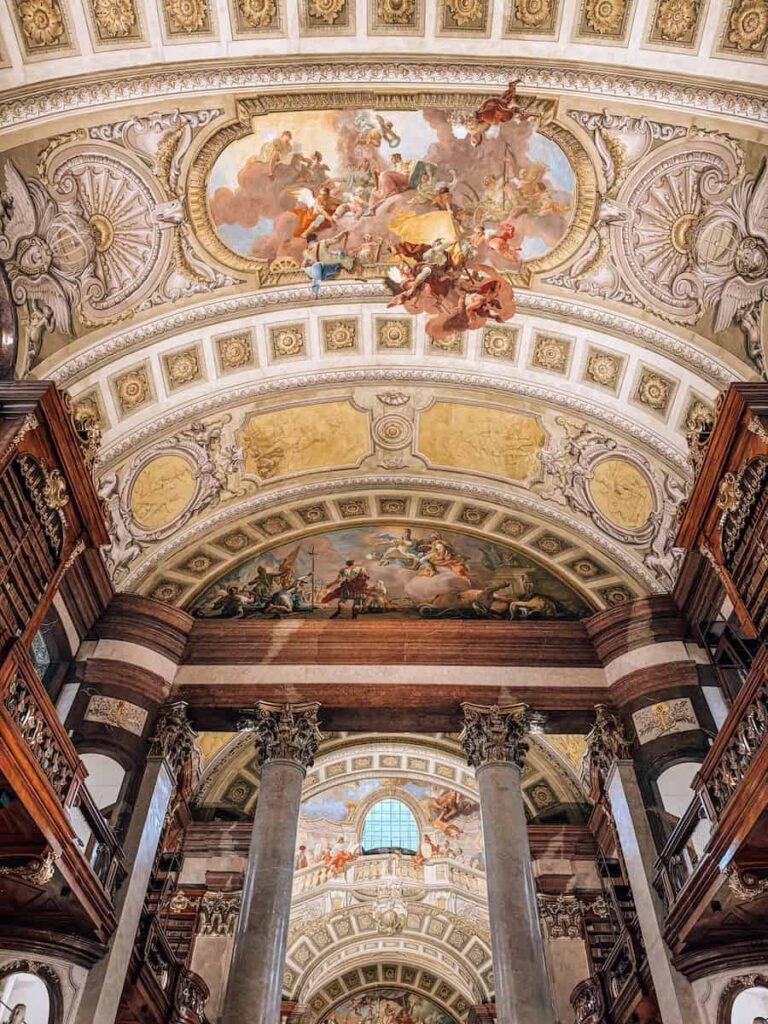 Interior view of the Austrian National Library in Vienna showcasing a grand Baroque hall with marble columns, ornate gold-trimmed balconies, and multiple ceiling frescoes depicting mythological scenes with vibrant figures and clouds.