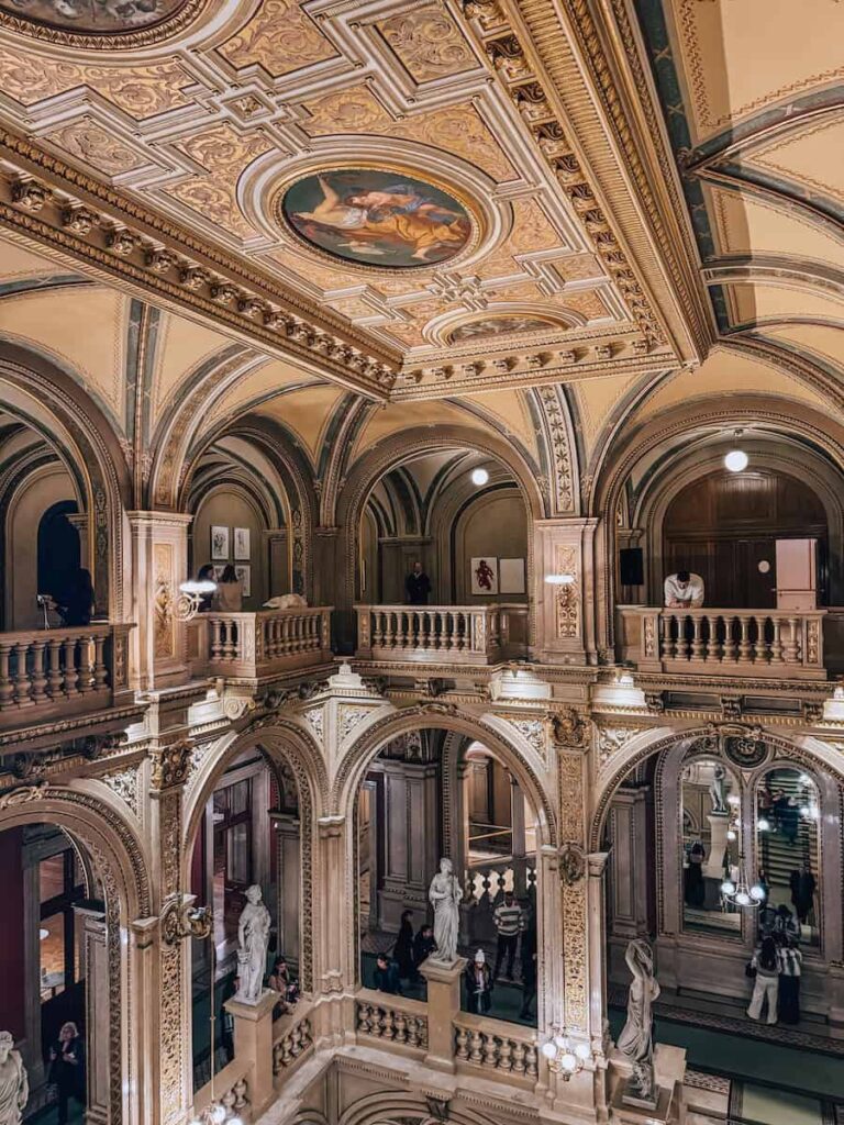 Interior view of the Vienna State Opera showcasing its richly decorated golden ceiling with frescoes, arched balconies, marble statues, and intricately detailed architecture. Visitors admire the opulent surroundings from various levels of the grand staircase.