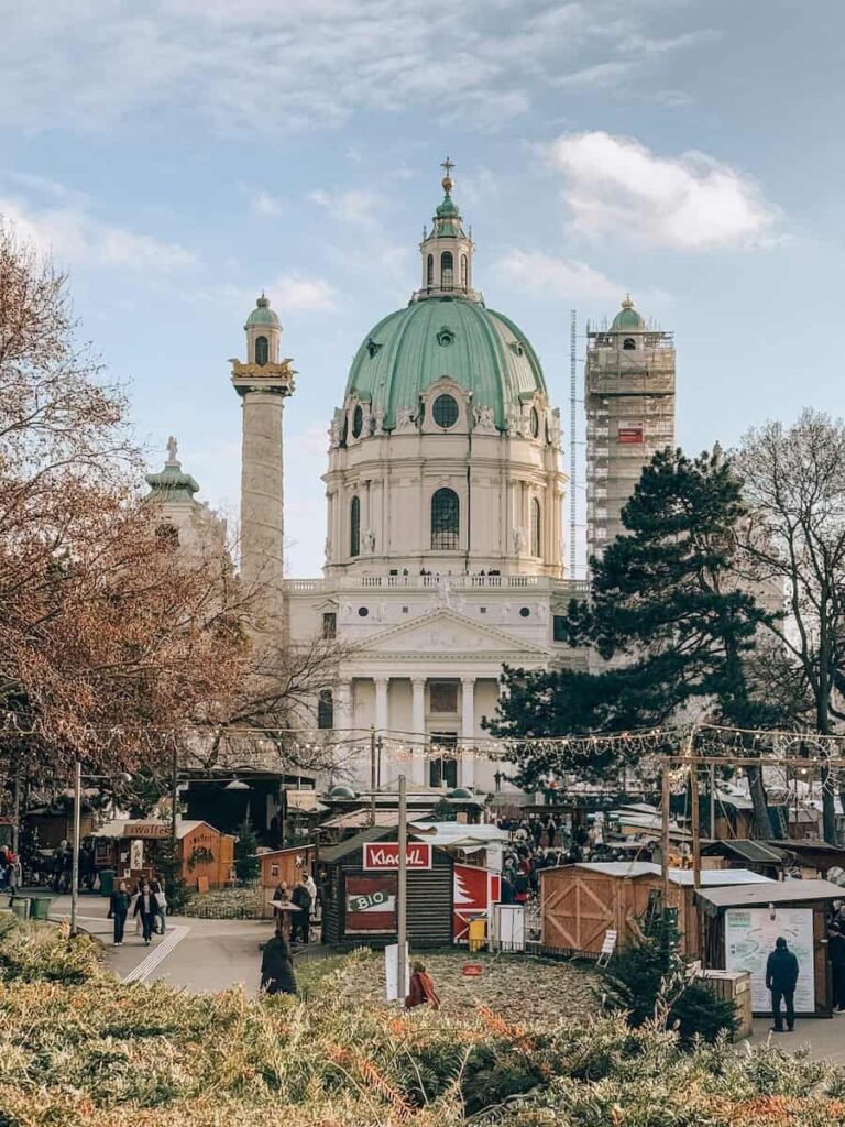 Stunning daytime view of Karlskirche (St. Charles's Church) in Vienna with its distinctive green dome and twin columns, surrounded by Christmas market stalls and visitors in the foreground, framed by winter trees and festive lights.