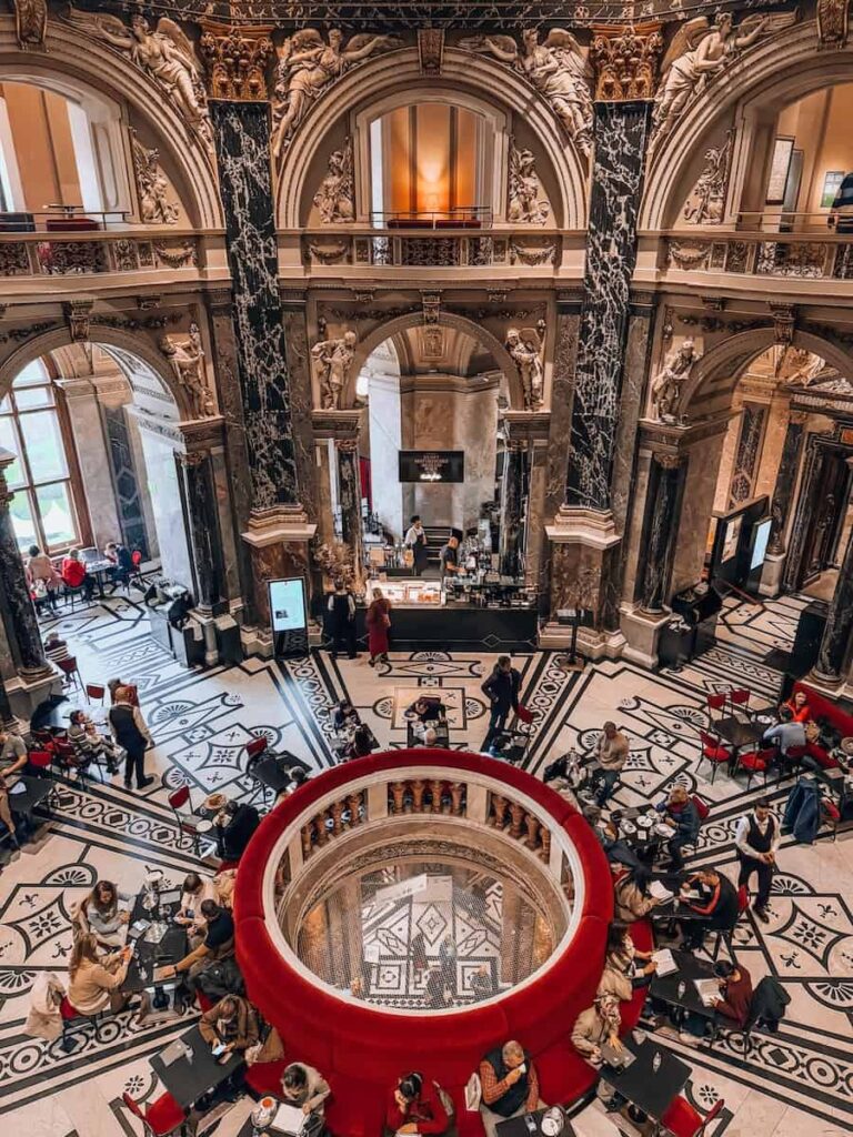 Overhead view of the café in the Cupola Hall of the Kunsthistorisches Museum in Vienna, featuring a grand circular opening with red velvet seating around it, black marble columns, intricate tile flooring, and guests enjoying coffee and pastries in a majestic, historic setting.