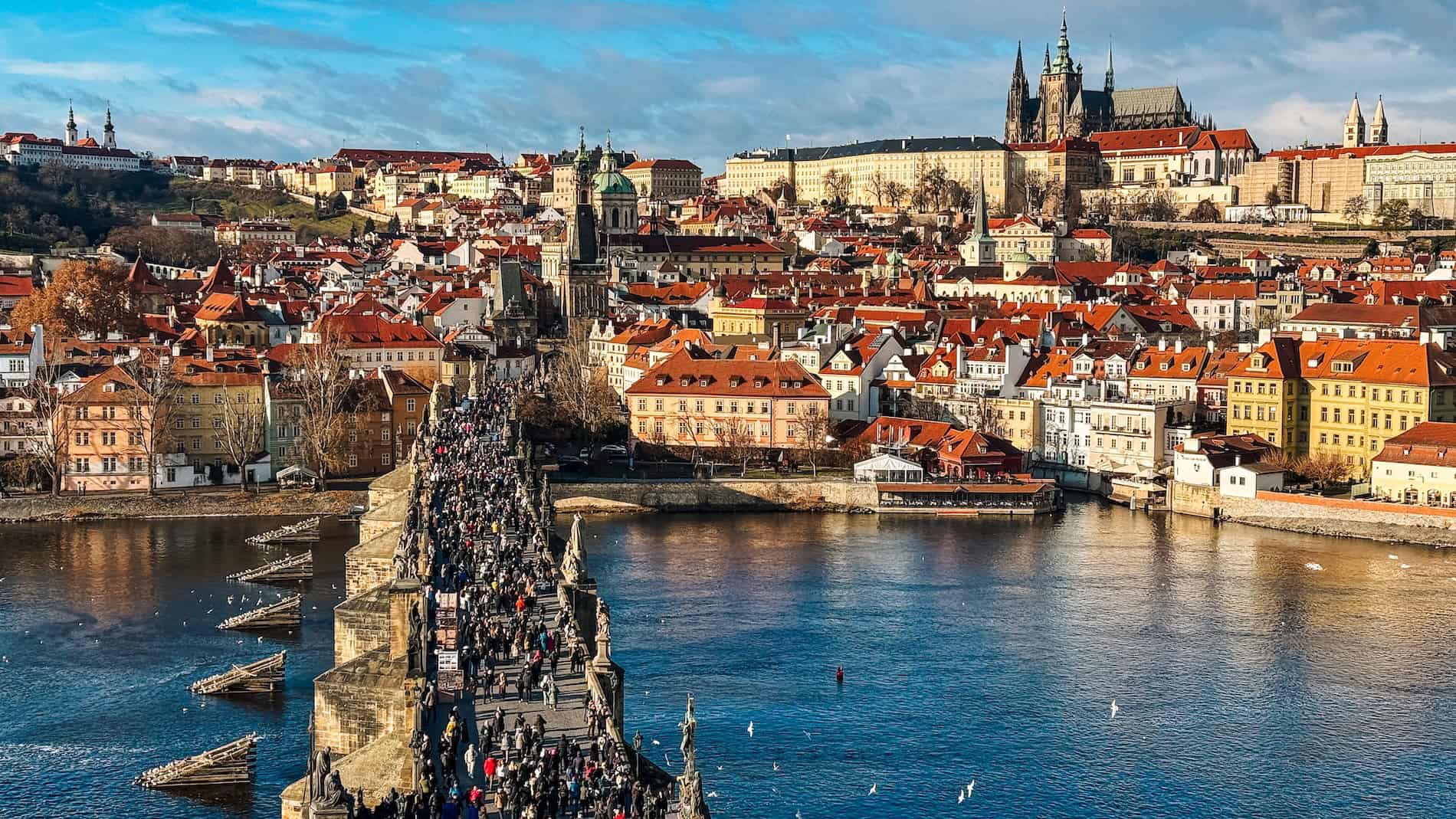 Panoramic view of Prague’s Charles Bridge crowded with pedestrians, spanning the Vltava River with colorful historic buildings and Prague Castle in the background under a bright blue sky