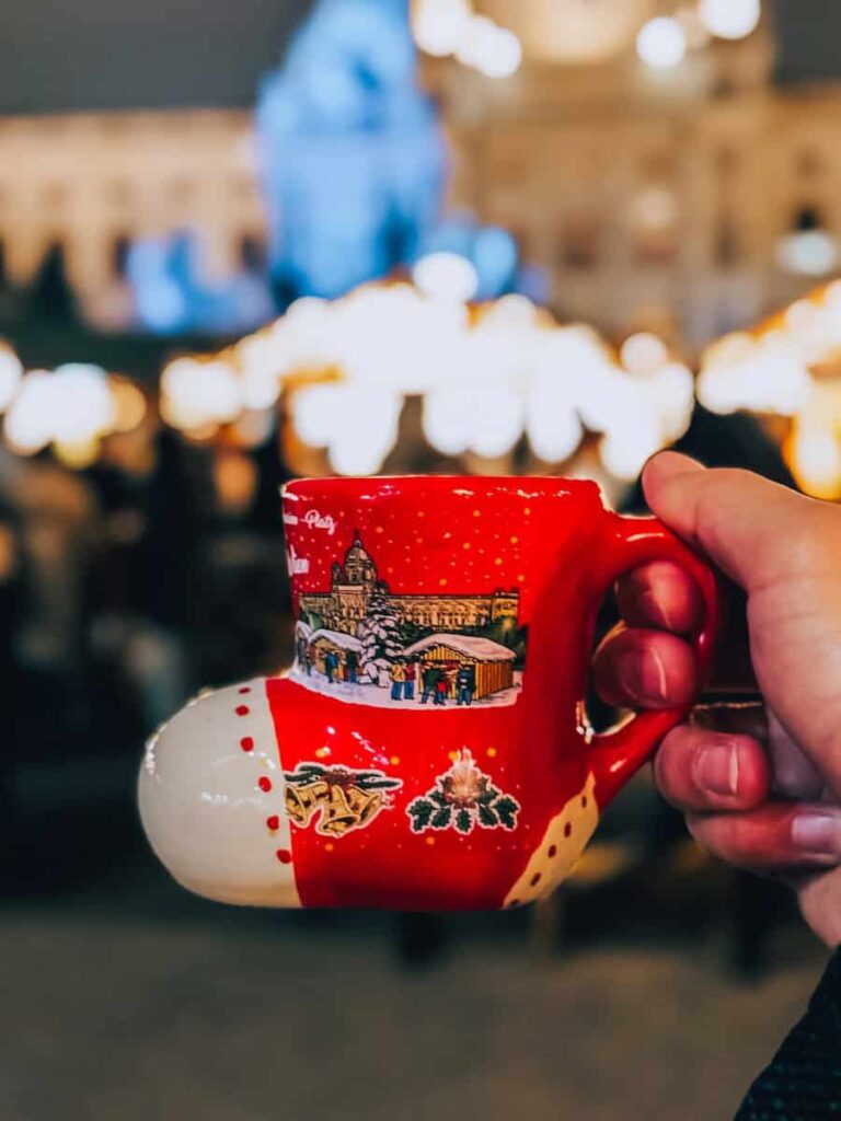 Close-up of a red boot-shaped Christmas market mug held in front of blurred festive lights and stalls at a Vienna Christmas market at night