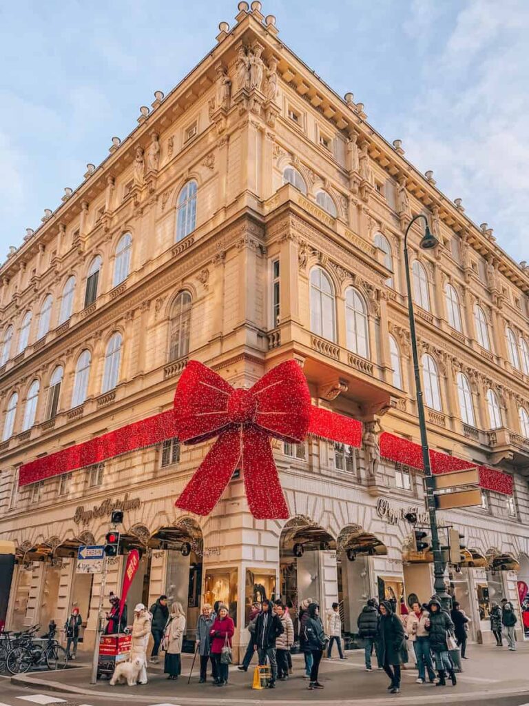 A grand corner building in Vienna wrapped in festive red ribbon with a giant glittery bow, bustling with holiday shoppers.