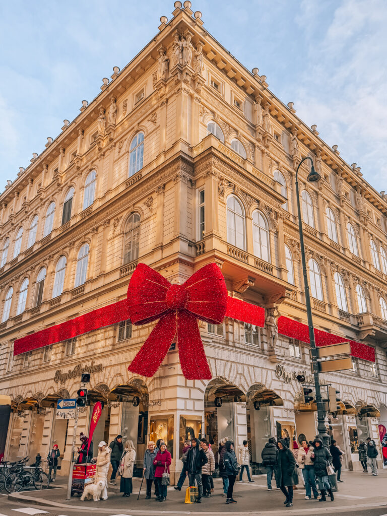 A grand corner building in Vienna wrapped in festive red ribbon with a giant glittery bow, bustling with holiday shoppers.