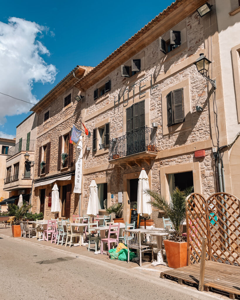 Colorful chairs and white umbrellas line the sidewalk outside a rustic stone building in Santanyí, Mallorca, under a bright blue sky.