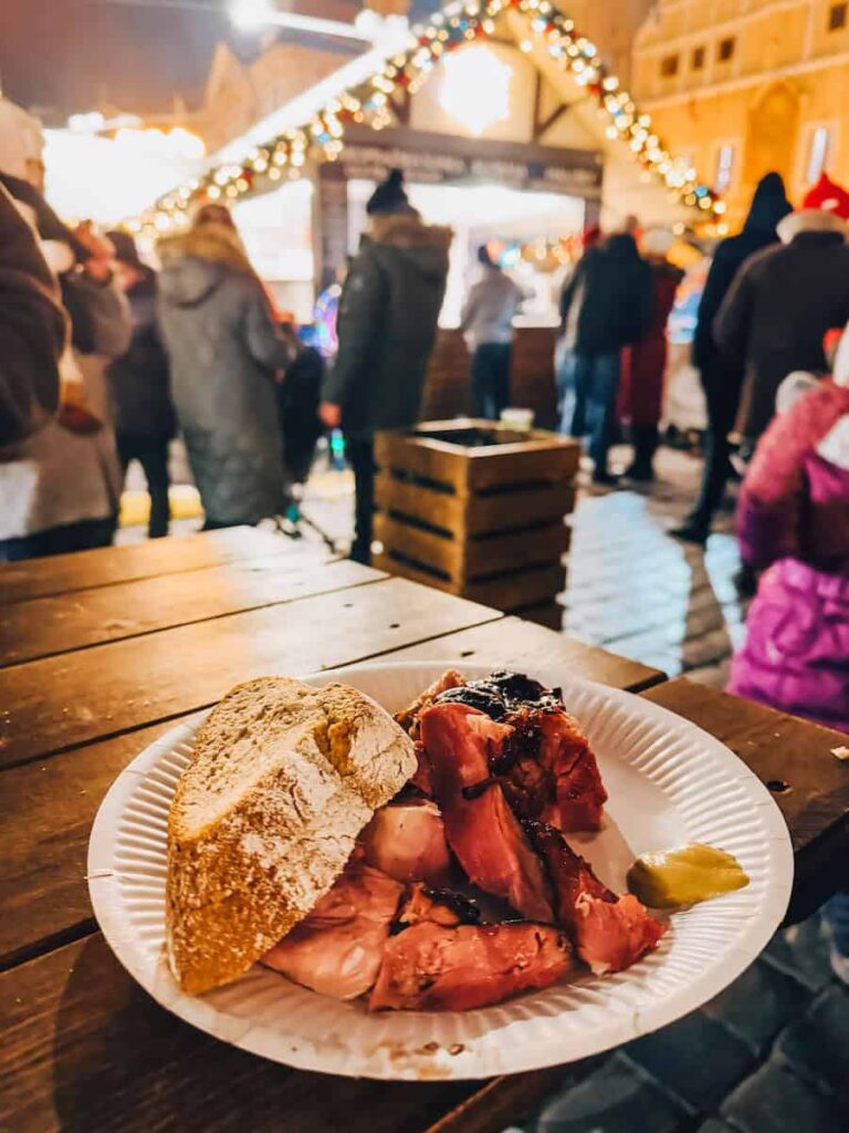 A close-up of a plate with sliced smoked meat, a crusty piece of bread, and a dollop of mustard, set on a wooden table at the Prague Christmas markets with festive lights and people in the background.