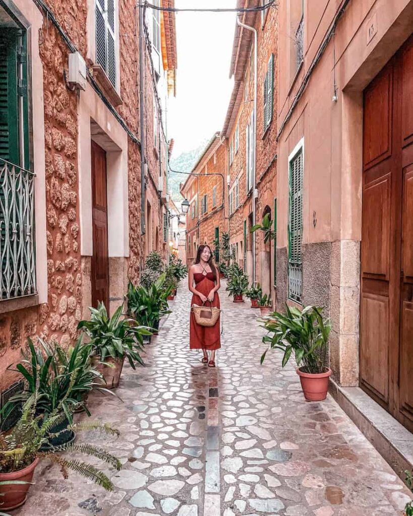 Woman in a rust-red dress walking along a narrow cobblestone street in Sóller, Mallorca, lined with potted green plants and traditional stone buildings with green shutters.