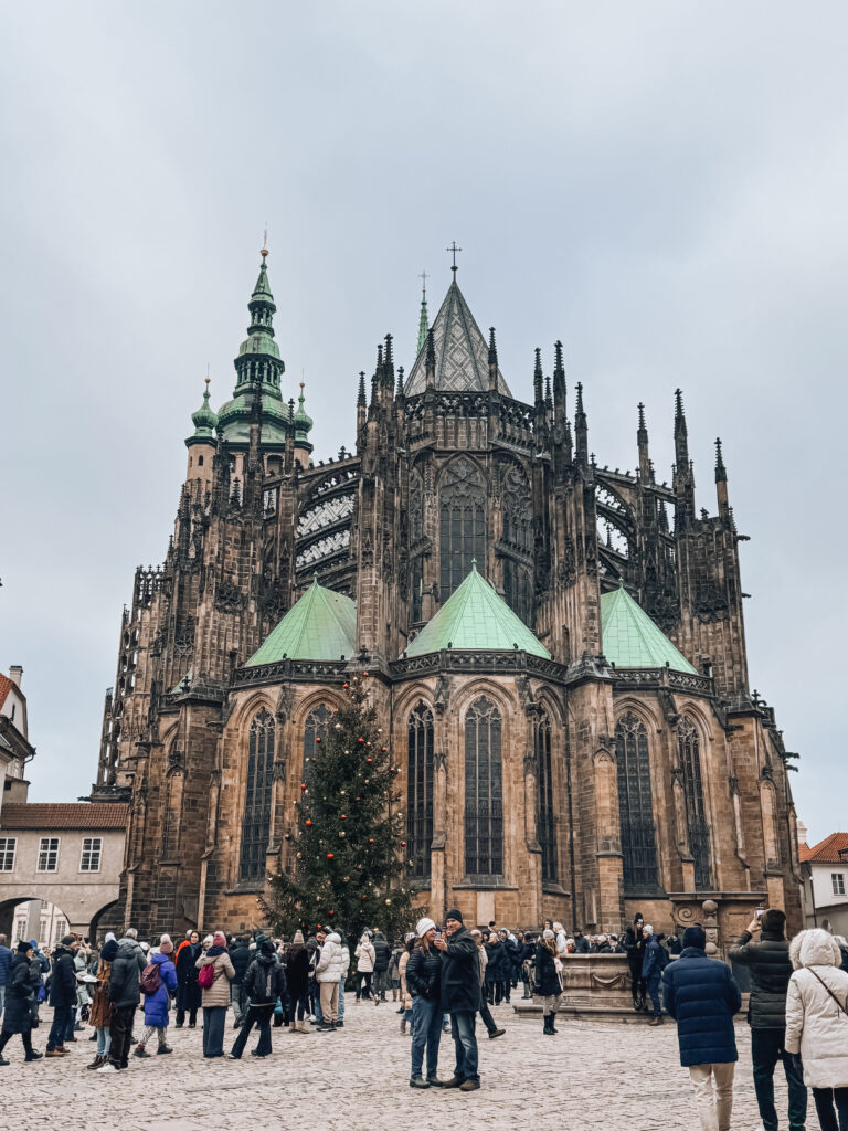 Crowds gathering in front of the rear façade of St. Vitus Cathedral in Prague, with a decorated Christmas tree standing in the courtyard during the winter holiday season