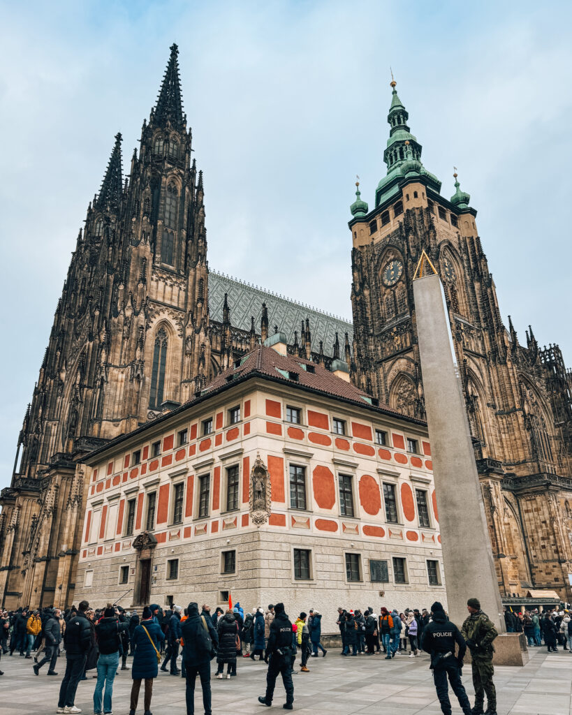 Visitors exploring the main courtyard of Prague Castle, with the Gothic spires and green domes of St. Vitus Cathedral towering behind the ornate red and white Old Royal Palace building