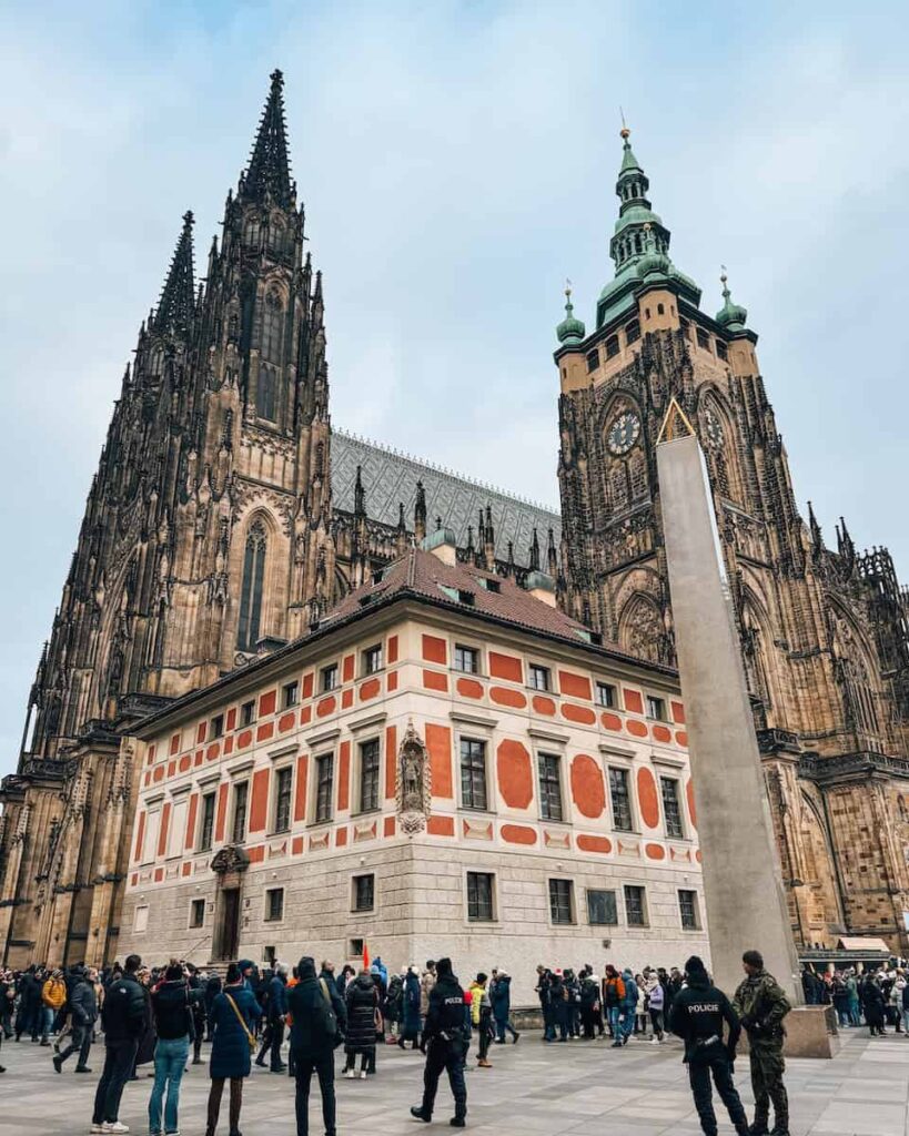 Visitors exploring the main courtyard of Prague Castle, with the Gothic spires and green domes of St. Vitus Cathedral towering behind the ornate red and white Old Royal Palace building