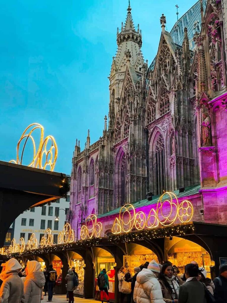 Crowds walking through the Stephansplatz Christmas Market in Vienna at dusk, with St. Stephen’s Cathedral illuminated in purple lights behind festive stalls and holiday decorations.