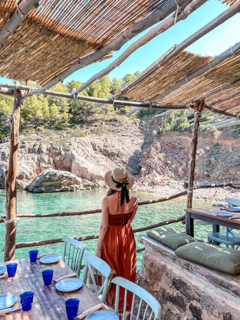 Woman in a rust-orange dress and straw hat standing on a rustic wooden terrace at a seaside restaurant in Cala Deià, Mallorca, overlooking turquoise waters and rocky cliffs.