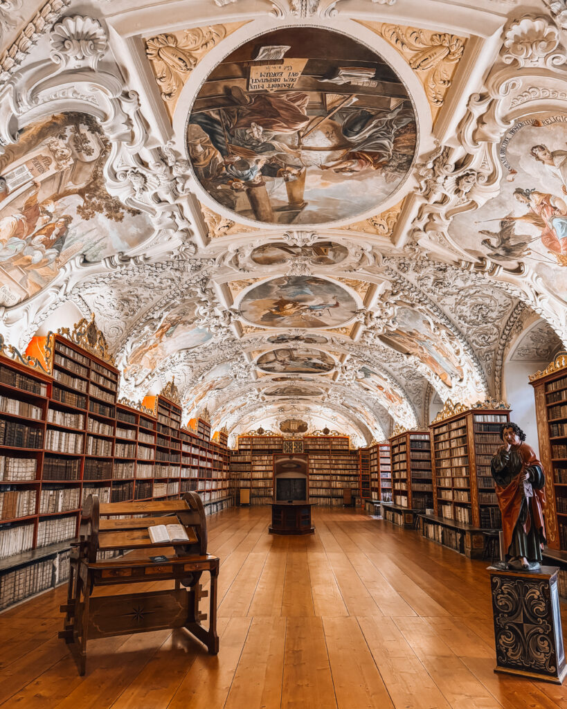 The Theological Hall at Strahov Monastery Library in Prague, featuring ornate Baroque ceiling frescoes, curved white stucco moldings, rows of ancient books, and polished wooden floors