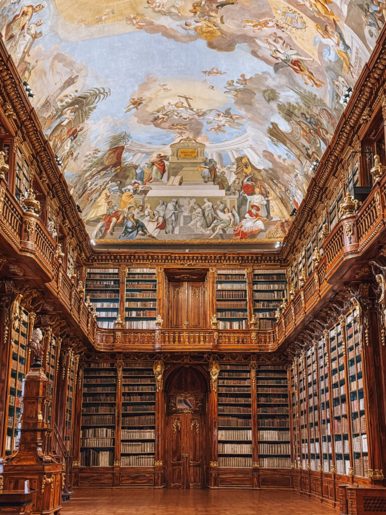 The Philosophical Hall at Strahov Monastery Library in Prague, with tall wooden bookshelves, richly carved details, and a soaring ceiling covered in elaborate Enlightenment-themed frescoes