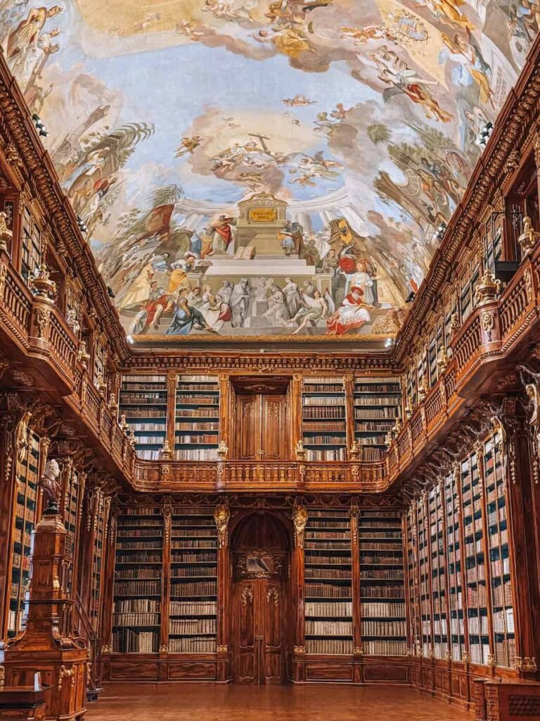 The Philosophical Hall at Strahov Monastery Library in Prague, with tall wooden bookshelves, richly carved details, and a soaring ceiling covered in elaborate Enlightenment-themed frescoes