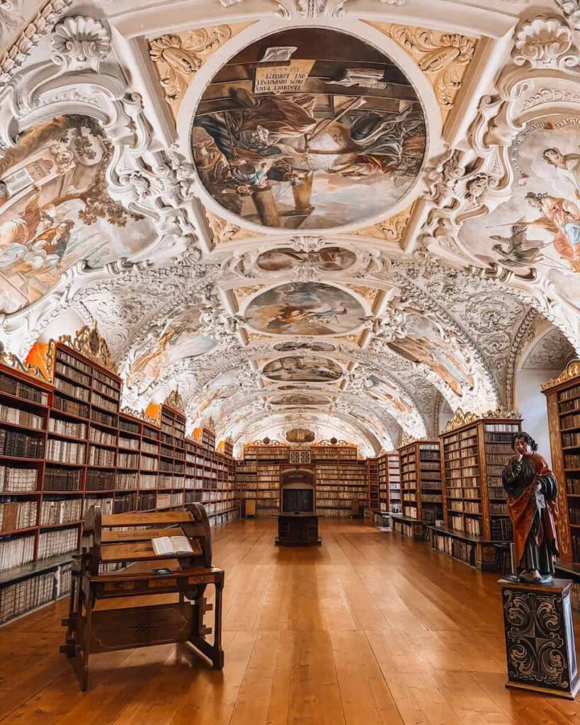 The Theological Hall at Strahov Monastery Library in Prague, featuring ornate Baroque ceiling frescoes, curved white stucco moldings, rows of ancient books, and polished wooden floors