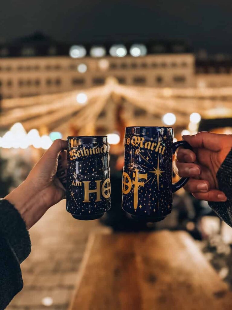Two people clinking festive blue mugs labeled "Weihnachtsmarkt am Hof" in front of golden Christmas lights at the Am Hof Christmas Market in Vienna.