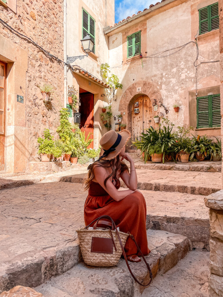 Traveler in a rust-colored dress and straw hat sits on rustic stone steps in Valldemossa, surrounded by historic buildings with green shutters.