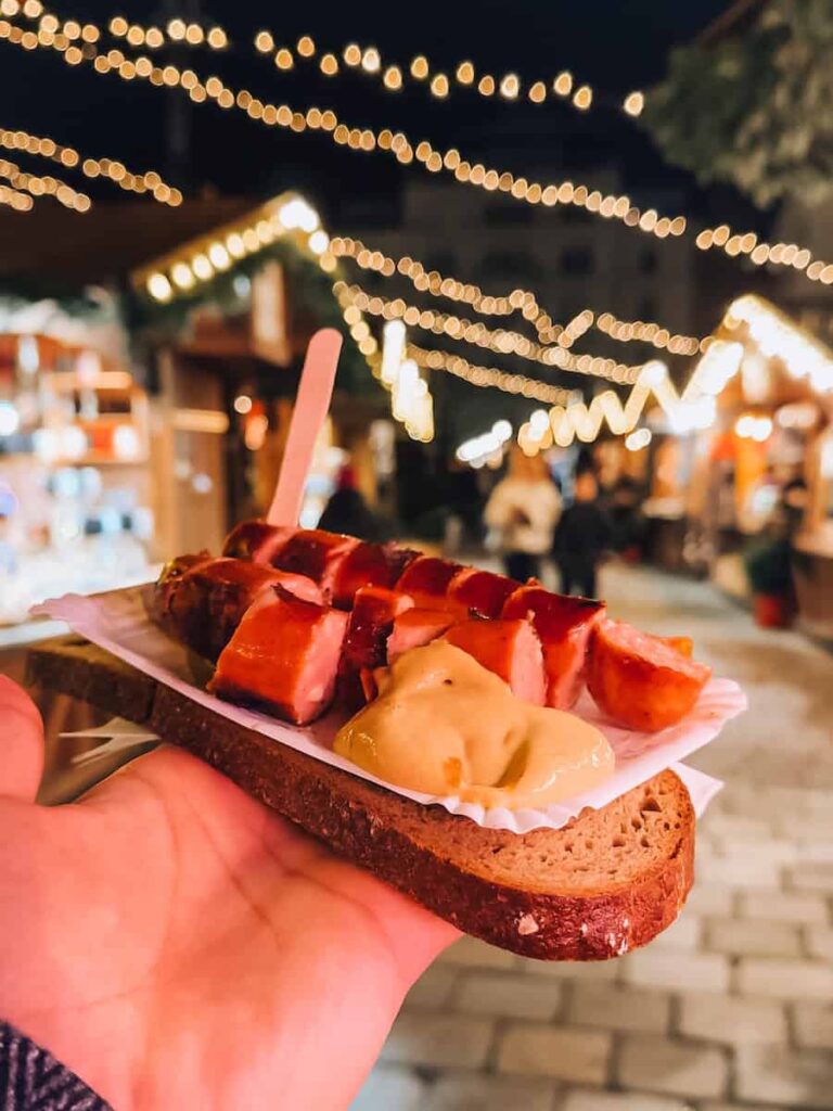 Traditional Austrian sausage sliced on a slice of rye bread and served with mustard, held in hand under glowing string lights at a Vienna Christmas market.