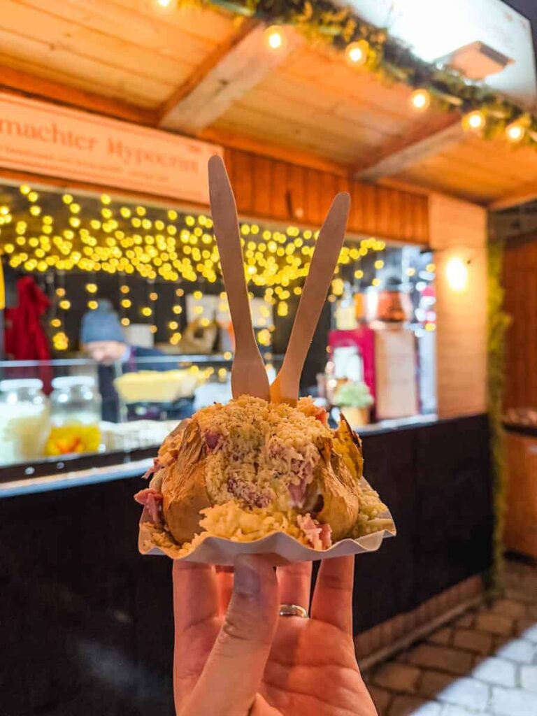 Close-up of a baked potato stuffed with chopped ham and shredded cheese, held up in front of a festive food stall at a Vienna Christmas market with warm fairy lights.
