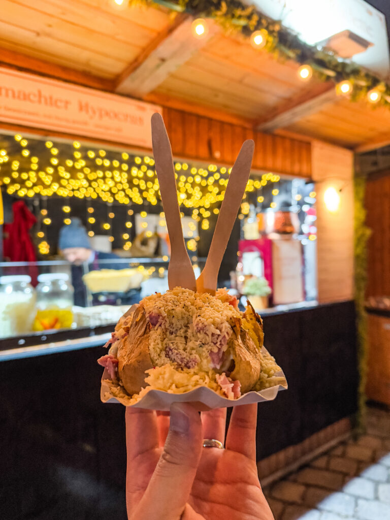 Close-up of a baked potato stuffed with chopped ham and shredded cheese, held up in front of a festive food stall at a Vienna Christmas market with warm fairy lights.