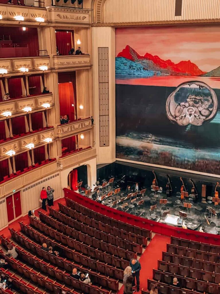 View of the main stage and orchestra pit inside the Vienna State Opera, with guests taking their seats. The grand interior features red velvet chairs, illuminated private balconies, and a large stage curtain displaying a modern artwork with abstract mountain and anatomical imagery.
