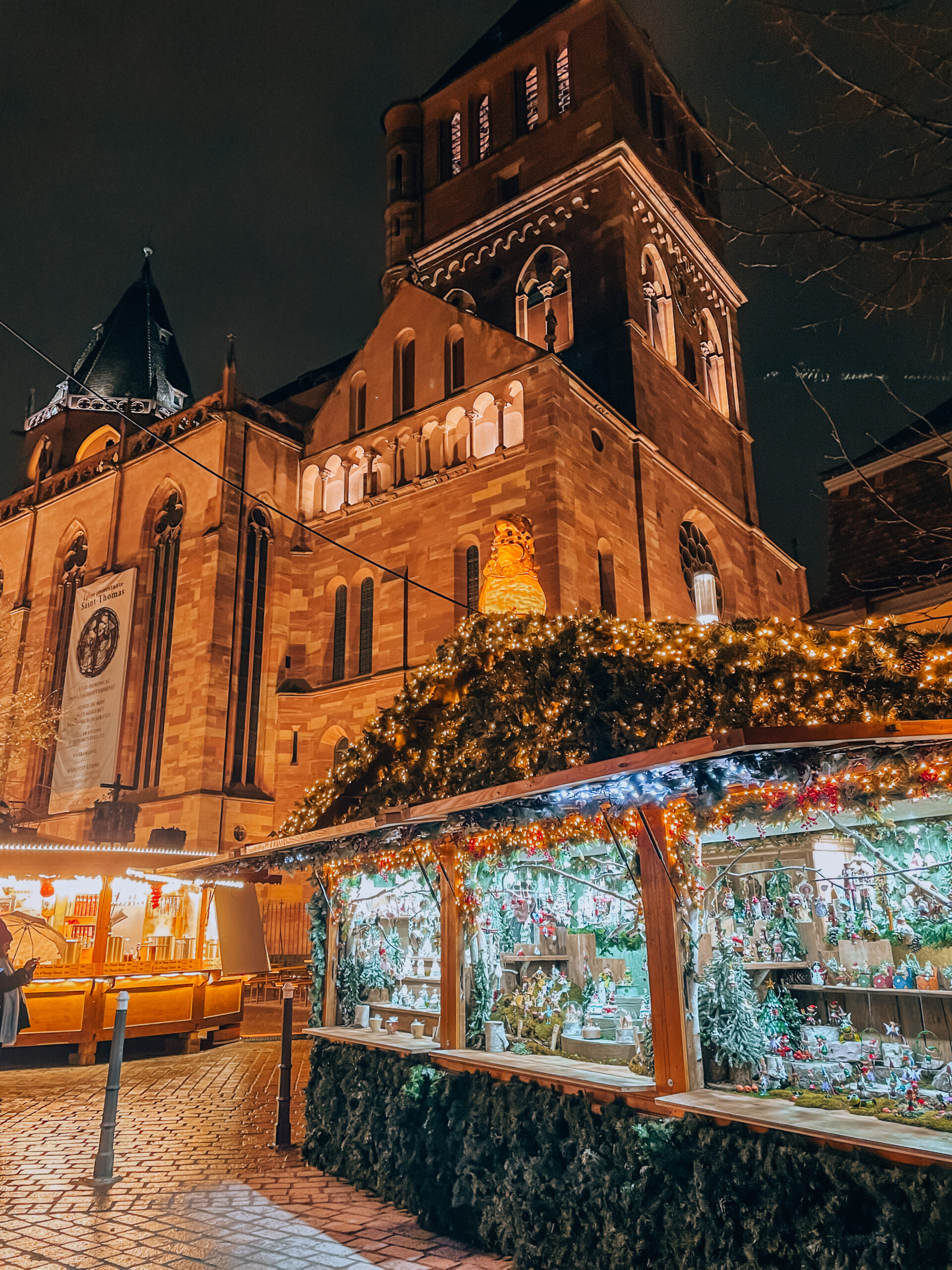 Wooden chalets decorated with garlands and lights at Strasbourg Christmas market with Saint Thomas Church glowing behind.