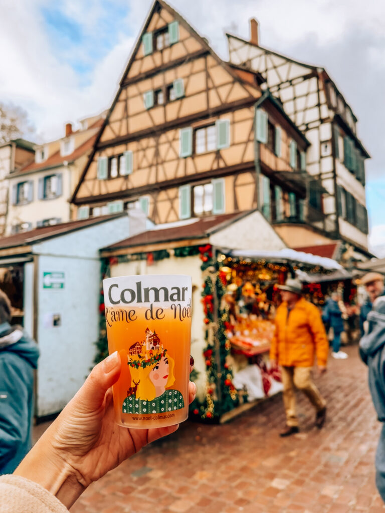Hand holding a festive Colmar Christmas market cup of hot cider with half timbered houses in the background.