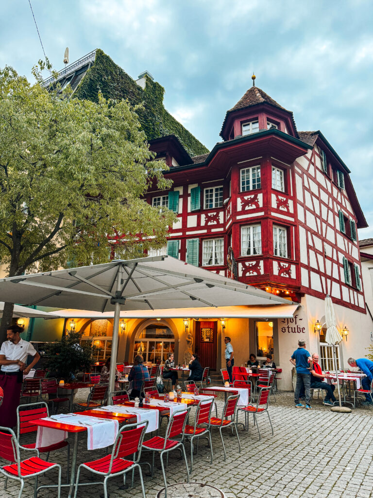 Red half-timbered building with ivy roof and outdoor dining terrace set with red chairs.