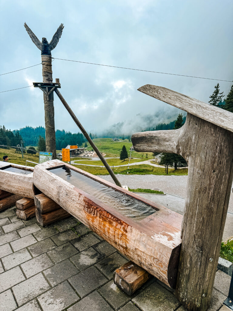 A rustic wooden water trough at Mount Pilatus with a tall carved eagle statue overlooking the misty mountain landscape.