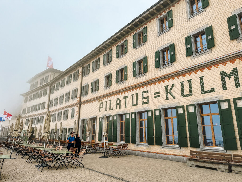The historic Pilatus Kulm hotel building with Swiss flags, outdoor seating, and fog rolling in around the mountain.