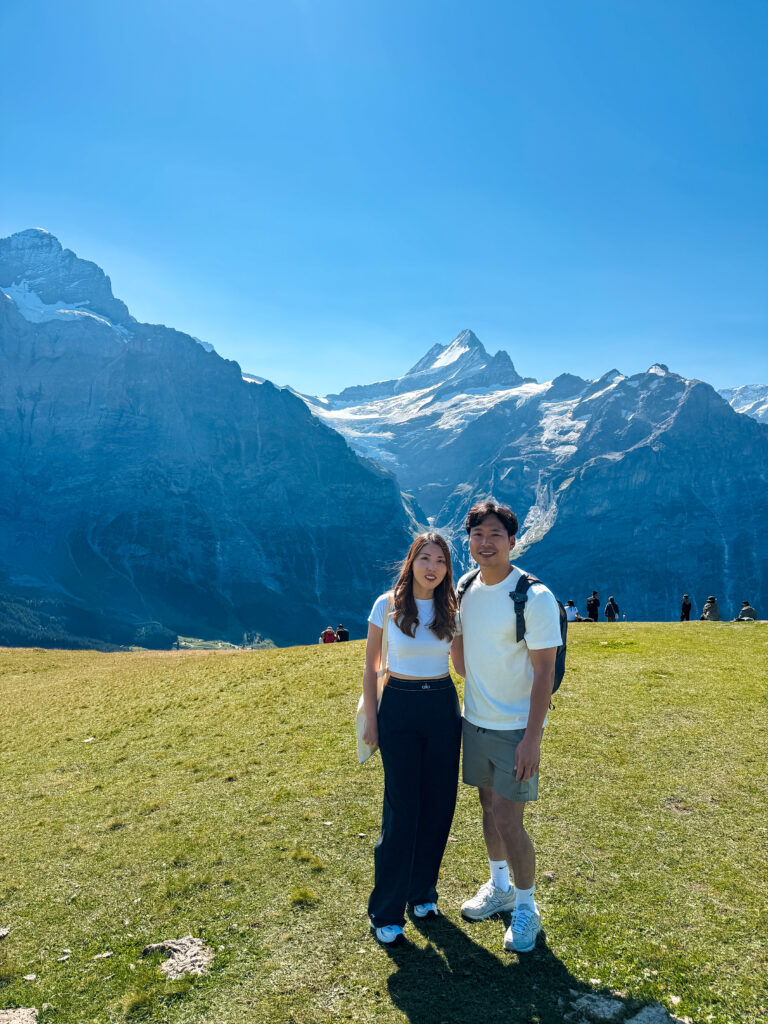 Couple standing on a grassy alpine meadow with the snow-capped Swiss Alps in the background on a clear blue day near Grindelwald.