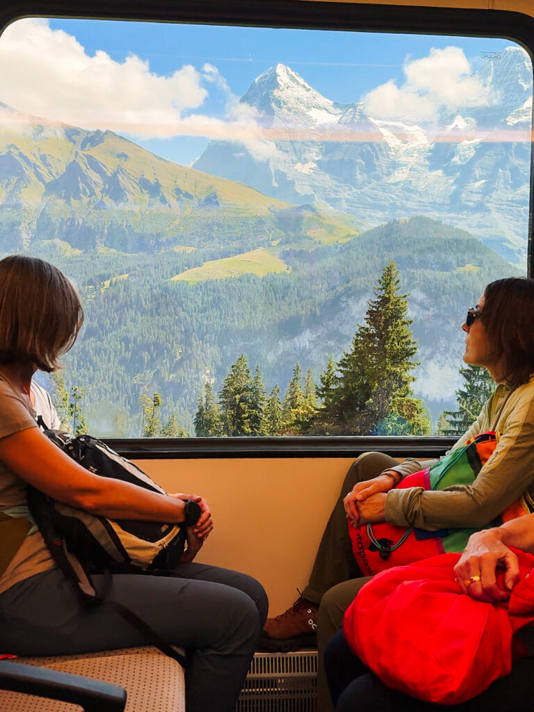 Train ride from Lauterbrunnen to Mürren with passengers gazing out the window at towering alpine peaks and rolling green hills.