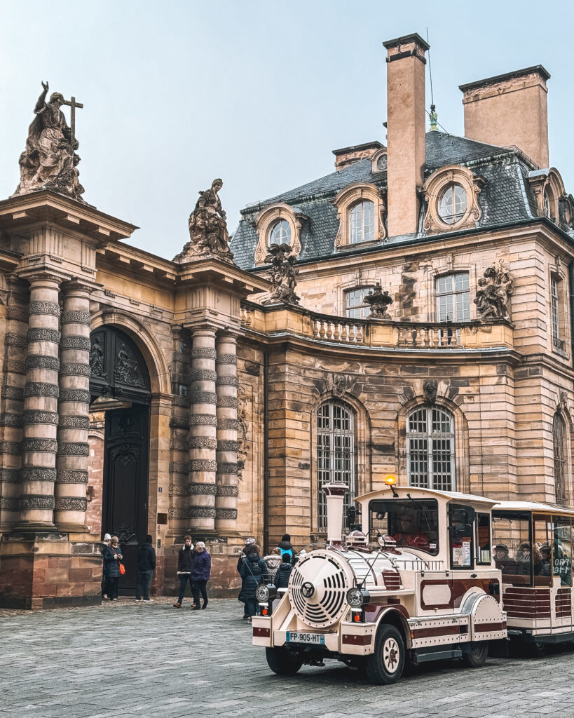 White and red tourist train parked in front of ornate historic architecture in Strasbourg.