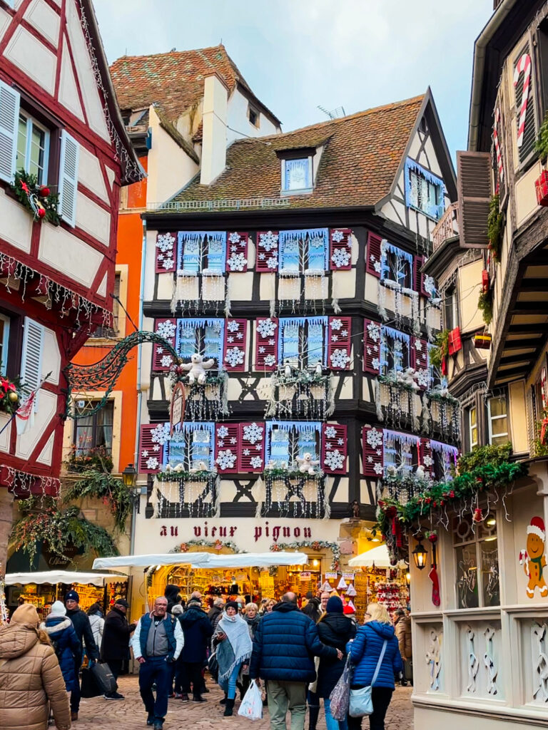 Busy street in Colmar with a half timbered building covered in snowflake shutters and festive decorations during Christmas markets, sign with au vieux pignon