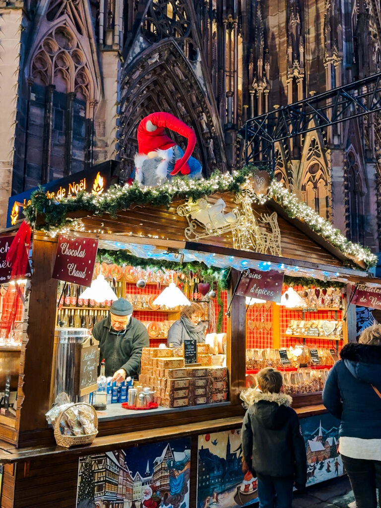 Wooden Christmas market stall decorated with garlands and lights selling Vin Chaud in front of Strasbourg Cathedral.