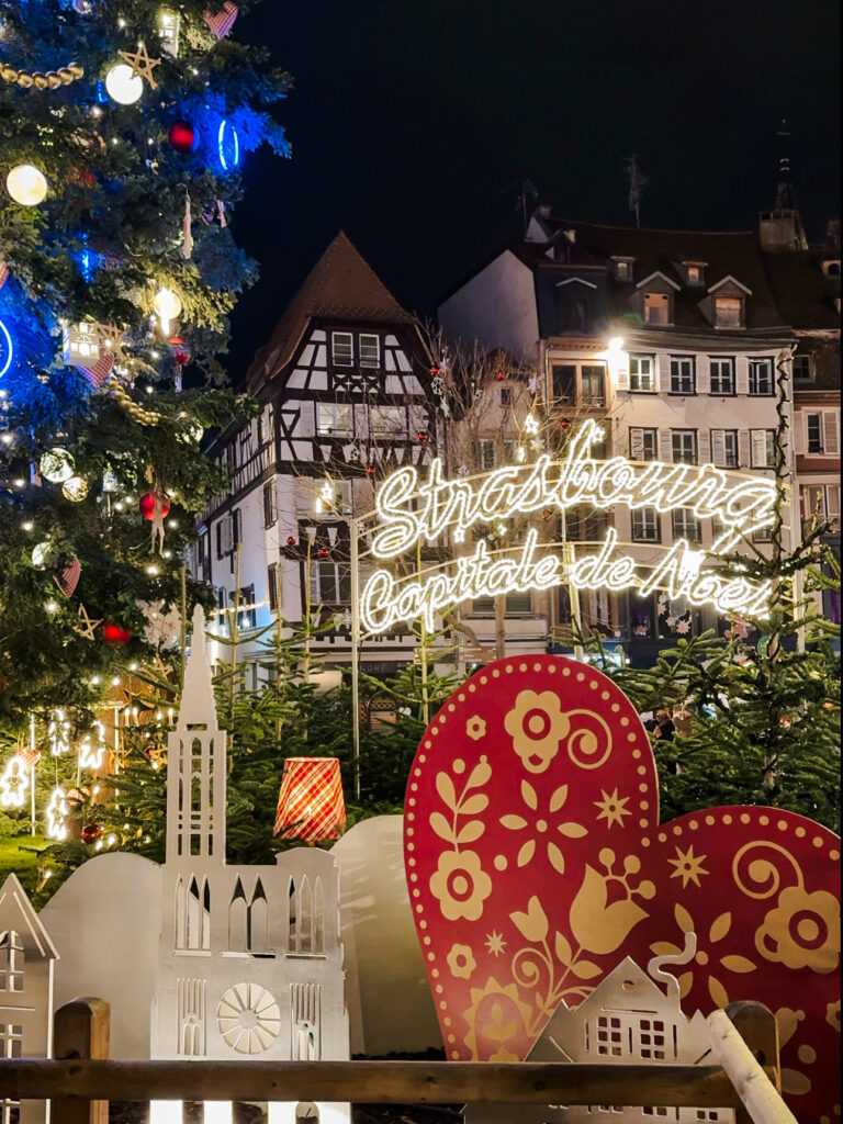 Festive Strasbourg Christmas display with glowing sign “Strasbourg Capitale de Noël” beside decorated trees.