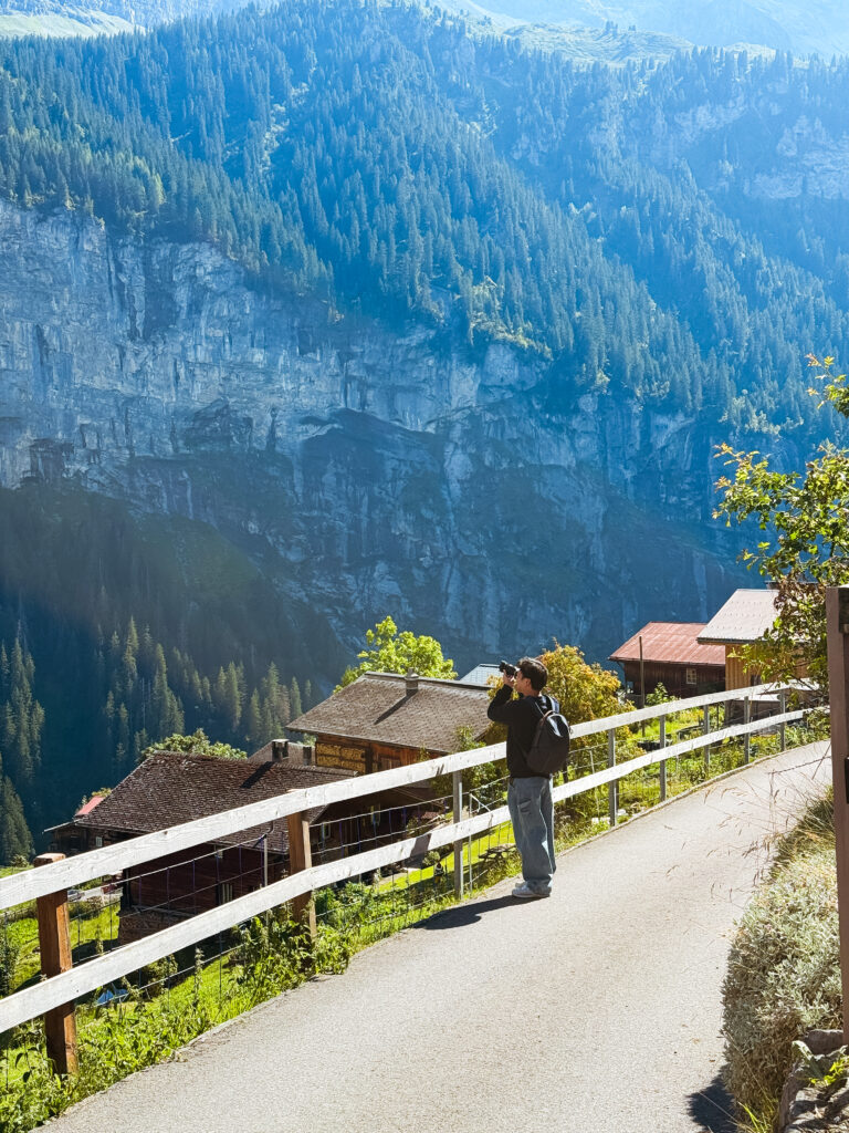 Traveler photographing the views in Gimmelwald from a hillside path overlooking alpine chalets and a dramatic forested gorge.