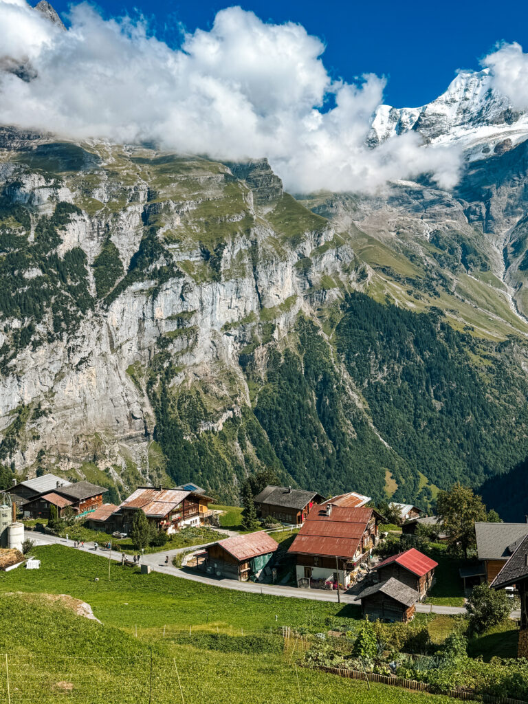Aerial view of Gimmelwald village with scattered wooden houses and barns set on a green slope against steep mountain cliffs.