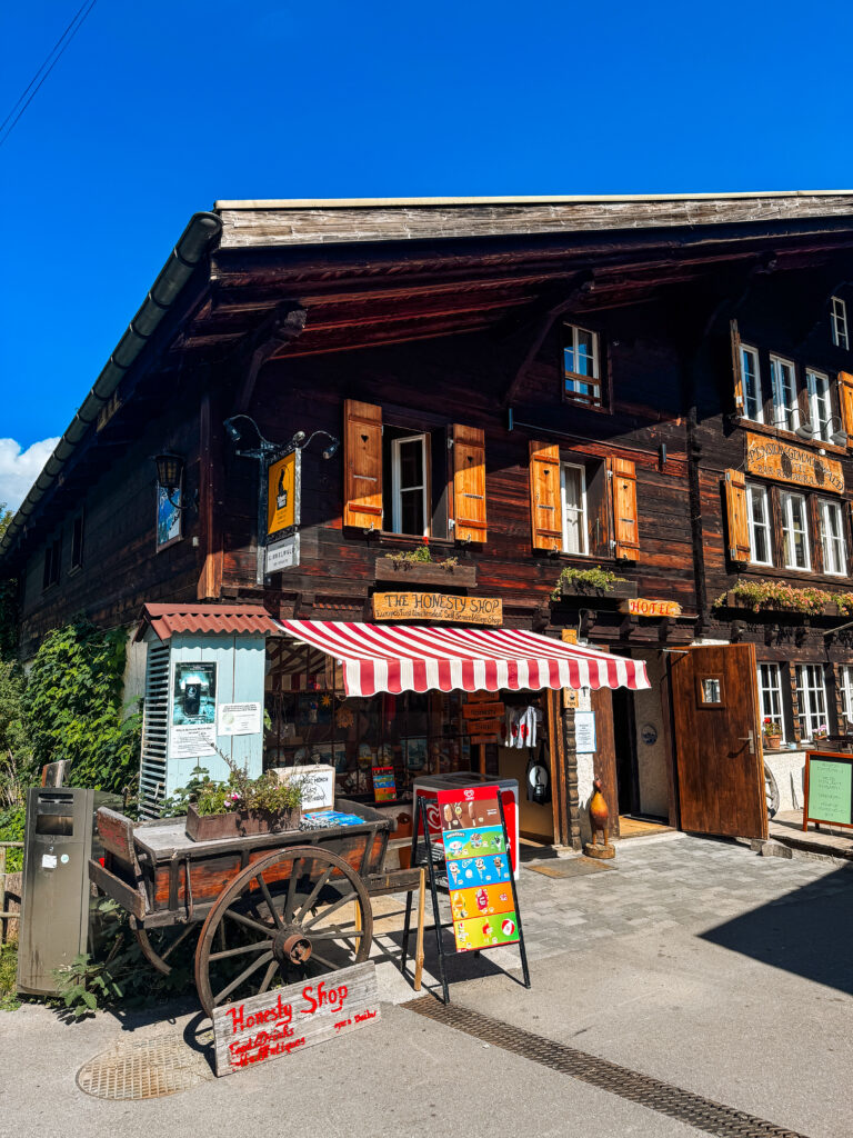 The Honesty Shop in Gimmelwald with a rustic wooden facade, striped awning, and wagon cart, set under a clear blue sky.